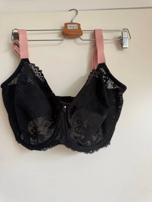Svart spets-bh med rosa axelband stl. 70 M - Snygg svart bh med spetsdetaljer och rosa axelband. Bh:n har bygel och dekorativ liten pärla framtill. Baksidan är slät med hakknäppning och sidorna har genomskinlig spets. Materialet är en mix av spets och satin.