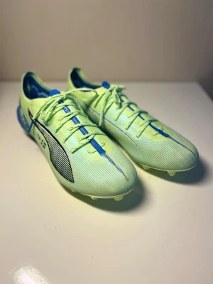 PUMA ULTRA 5 ULTIMATE FG LIGHTS OUT - GRÖN/PUMA WHITE/BLÅ - Säljer ett par snygga ljusgröna fotbollsskor från Puma med blå och svarta detaljer samt svensk flagga på sidan. Skorna har snörning, låg profil och är tillverkade i syntetmaterial för bästa grepp på planen.