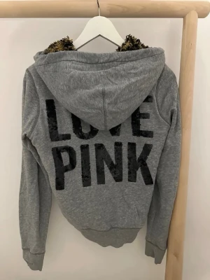 Grå hoodie från Victoria's Secret PINK - Grå zip-hoodie från Victoria's Secret PINK med stora svarta paljettbokstäver 'LOVE PINK' på ryggen. Mjuk huva med leopardmönstrat foder samt fickor fram. 