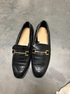 Svarta loafers med krokodilstruktur H&M - Stilrena svarta loafers från H&M med krokodilpräglat mönster och guldfärgade metalldetaljer framtill. Skorna har en klassisk form och låg klack, perfekta för en trendig och chic look. Snyggt konstläder som ger en lyxig känsla.
