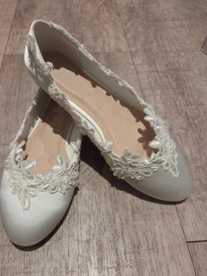 Satinskor i Ballerinamodell med spetsdetaljer st 37 - Satinskor i Ballerinamodell med spets och broderade blommor samt pärldetaljer runt kanten. Färg Ivory Skorna har rund tå och är tillverkade i lyxig  satin. Perfekta för dig som vill ha något extra till outfiten.