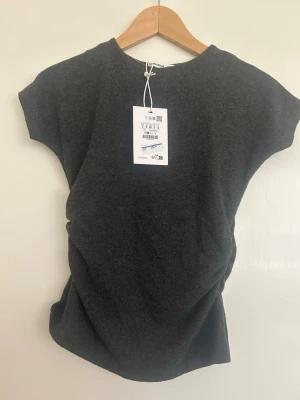 Mörkgrå topp med scrunch-detalj - Stilren mörkgrå topp från Pull&Bear med scrunch-detalj på sidan som framhäver midjan och ger en mer figurnära och smickrande passform. Supermjuk och bekväm i materialet. Lätt att matcha till både jeans, kjol eller kostymbyxor. Ny med prislapp kvar