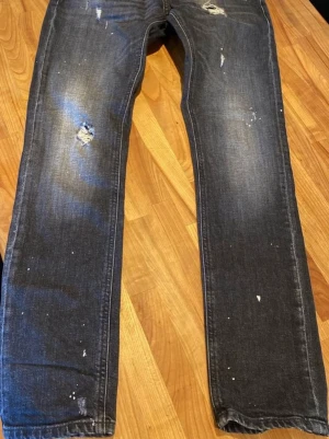 Mörkgrå slitna jeans med raka ben - Säljer ett par mörkgrå jeans Dondup George skinny fit med raka ben och slitningar på låret och knät. Jeansen har en tvättad look med små vita färgstänk och är tillverkade i klassisk denim. Perfekta för en avslappnad och trendig stil. I mycket gott skick, sparsamt använda. Ca 32 i längd.