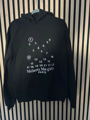 Maison Margiela svart hoodie - Svart hoodie från Maison Margiela med ikoniskt vitt tryck av siffror och logotyp på bröstet. Klassisk huva och ribbade muddar. Perfekt för dig som gillar stilrena designer och vill sticka ut med ett exklusivt plagg.
