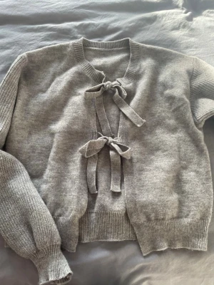 Grå stickad kofta med knytband - Superfin grå stickad kofta med två knytband framtill. Koftan har långärmade ärmar och ribbade muddar vid hals, ärmslut och nederkant. Perfekt att slänga över en topp för en chill och snygg look.