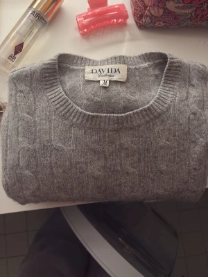 Grå cashmere tröja från Davida - Säljer en grå kabelstickad tröja från Davida i storlek M. Tröjan är gjord av mjuk cashmere och har rund halsringning samt ribbade muddar. Perfekt för att hålla sig varm och snygg under kyliga dagar😊😊