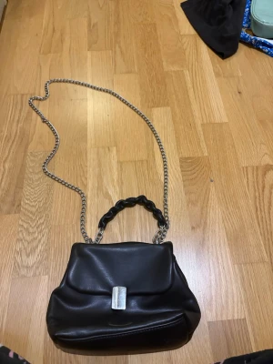 Svart handväska med kedjerem - Stilren svart handväska i slätt konstläder med silverfärgad kedjerem och ett flätat handtag. Väskan har ett lock med metallspänne och ett innerfack med dragkedja för småsaker. Perfekt accessoar för att lyfta din outfit.