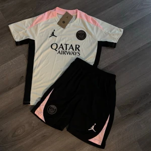 PSG x Jordan fotbollskit svart/rosa-M/L - Snyggt PSG x Jordan fotbollskit med tröja och shorts. Tröjan är vit med svarta och rosa detaljer, PSG-logga och Jordan-logga på bröstet samt Qatar Airways-tryck. Shortsen är svarta med rosa detaljer och PSG/Jordan-logga. Perfekt för träning eller match.