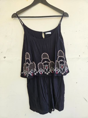 Svart broderad jumpsuit med tunna axelband - Svart jumpsuit från Cubus med tunna axelband och broderade detaljer med pärlor framtill. Fickor på båda sidor. Lite sliten vid knäppningen i ryggen, annars väldigt fint skick. 