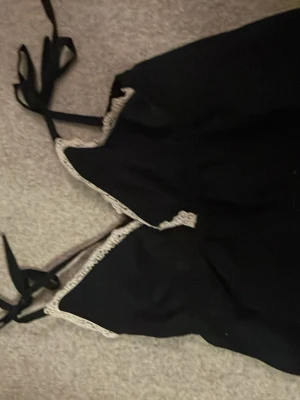 Svart linne med spets från RYVLS - Säljer ett svart linne från RYVLS med tunna axelband som knyts och vit spetsdetalj längs urringningen. Materialet känns mjukt och följsamt, perfekt för mysiga sommarkvällar. Enkel och stilren design med en feminin touch.