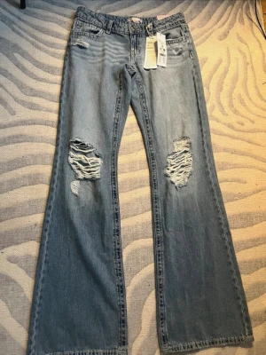 Ljusblå bootcut jeans med slitningar - Säljer ett par ljusblå bootcut jeans med stora slitningar på båda knäna. Jeansen har klassisk femficksmodell, låg midja och vida ben. Materialet är denim i bomull och de har en avslappnad vintagekänsla. Perfekta för dig som gillar en trendig och avslappnad stil.