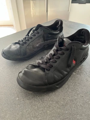 Svarta sneakers från Polo Ralph Lauren - Svarta låga sneakers i läder från Polo Ralph Lauren med svart sula och svarta skosnören. Det finns tydligt slitage på insidan vid hälen och några veck i materialet (se bilder), därför säljer jag dem billigare. Bra som vardagsskor eller extrasneakers.