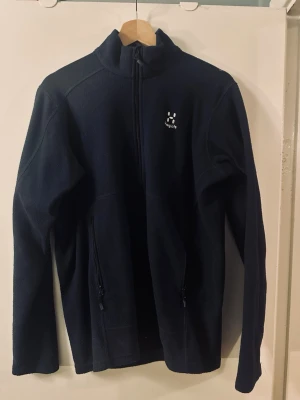 Marinblått fleecejacka från Haglöfs - Mysig svart fleecejacka från Haglöfs med hel dragkedja framtill och två sidofickor med dragkedja. Jackan har hög krage och lång ärm, perfekt för kyliga dagar. Klassisk och enkel design med Haglöfs-logga på bröstet.