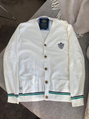 Stickad kofta från Morris med brodyr  - Vit cardigan från Morris med broderad logga på bröstet och snygga knappar framtill. Tröjan har ribbade muddar med gröna och svarta ränder, två fickor och förstärkta armbågar. Materialet är mjukt och tunt, perfekt för lager-på-lager.