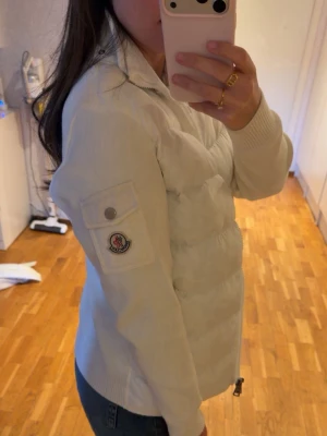 Vit dunjacka från Moncler - Snygg vit dunjacka från Moncler med stickade ärmar och ribbade muddar. Jackan har dragkedja framtill, ficka med knapp på ärmen och Moncler-logga. Perfekt för kalla och varma dagar och riktigt trendig look.