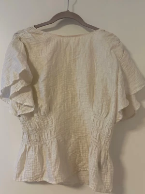 Beige blus med volangärmar - Luftig vit blus med korta volangärmar och smockad midja. Tillverkad i ett mjukt bomullsliknande material som ger en avslappnad och trendig look. Perfekt att styla med jeans eller kjol.