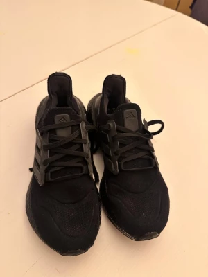 Svarta Adidas Ultraboost sneakers - Säljer ett par helsvarta Adidas Ultraboost sneakers med klassisk mesh- och textilovandel, svarta snören och ikoniska tre ränder på sidan. Skorna har en chunky sula och är perfekta för en sportig och stilren look.