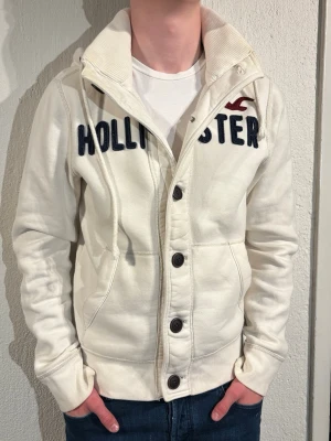 Vit hoodie från Hollister med knappar - Säljer en vit hoodie från Hollister med stora mörkblå bokstäver och röd logga på bröstet. Hoodien har knäppning med stora knappar, dragsko i huvan och två fickor framtill. Tillverkad i mjukt bomullsmaterial och har ribbade muddar vid ärmslut och nederkant.