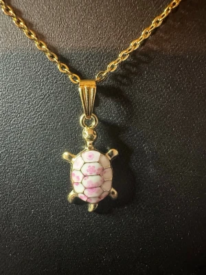 Guldhalsband med sköldpaddshänge - Unikt halsband i guldfärgad metall med ett sköldpaddshänge. Hänget har ett skal i vitt och rosa med små blommor, vilket ger en söt och lekfull känsla. Perfekt för dig som gillar djurmotiv och vill ha något som sticker ut.