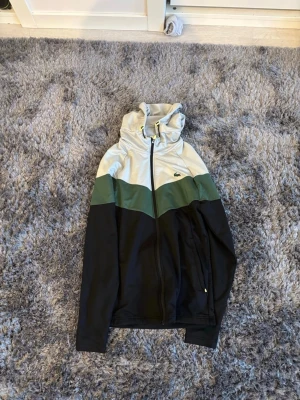 Vindjacka från Lacoste Sport, färgblock - Snygg vindjacka från Lacoste Sport med färgblock i svart, grönt och ljusgrått. Jackan har huva med dragsko och dragkedja framtill. Ikoniska Lacoste-loggan på bröstet och praktiska fickor med dragkedja. Perfekt för en sportig och trendig look.