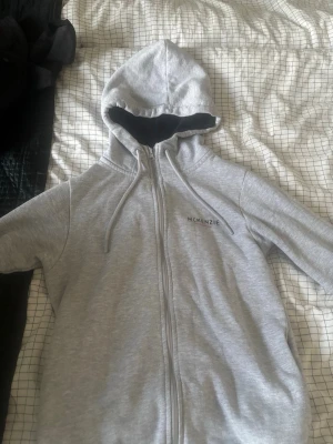 Grå zip hoodie från McKenzie - Ljusgrå hoodie med dragkedja från McKenzie. Klassisk modell med huva, dragsnören och svart innerfoder i huvan. Diskret logga på bröstet och mjukt material som känns skönt mot huden. Perfekt för en avslappnad och stilren look.