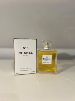 Chanel No 5 Eau de Parfum 100 ml - Klassisk Chanel No 5 Eau de Parfum i elegant, fyrkantig glasflaska med genomskinlig kork och vit etikett. Doften är ikonisk och tidlös, och flaskan rymmer 100 ml. Kommer med originalförpackning i stilren vit kartong med svart text.