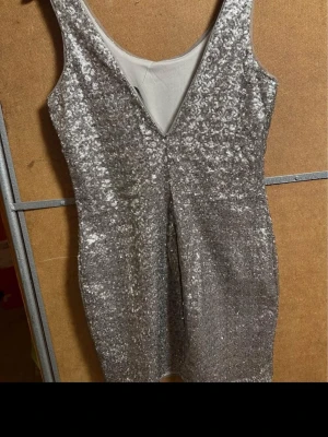 Glittrig silver kortklänning - Snygg glittrig silverfärgad kortklänning med djup v-ringning och ärmlös design. Klänningen har en figurnära passform och är täckt av paljetter som ger en festlig look. Perfekt för dig som vill sticka ut på nästa event.