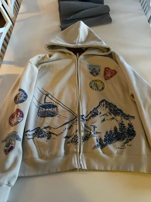Beige hoodie med bergstryck och patches - Säljer en beige hoodie med dragkedja från MAN. Den har blått tryck av berg och linbana på framsidan samt flera färgglada patches på bröstet och ena ärmen. Hoodien har huva och två fickor framtill. Perfekt för dig som gillar outdoor-vibbar och unik stil.