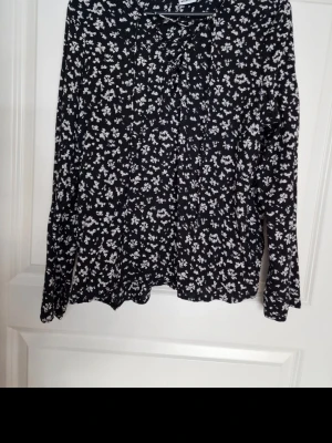 Svart blommig blus med snörning - Snygg svart blus med vita småblommiga mönster och dekorativ snörning vid halsen. Blusen har lång ärm och ett lätt, luftigt tyg som ger en avslappnad men trendig look. Perfekt att matcha med jeans eller kjol för en cool vardagsstil.