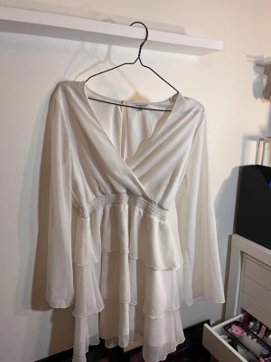 Vit klänning M/L(42) - Säljer en vit kortklänning med omlottliv och långa ärmar. Klänningen har flera lager volanger och smock i midjan för en snygg passform. Använd en gång, skulle säga att den är storlek M / L och inte 42
