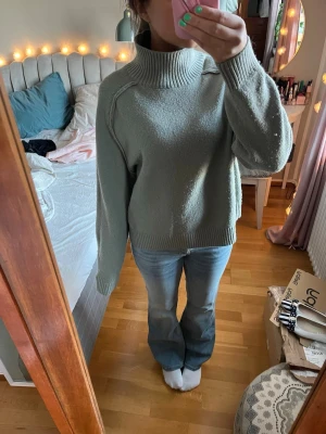 Grågrön stickad polotröja - Säljer en stickad tröja i grågrönt med hög polokrage och långa ärmar. Tröjan har ribbade muddar vid ärmslut och nederkant. 