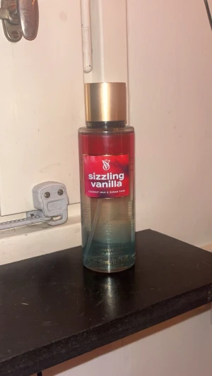 Victoria's Secret Sizzling Vanilla mist - Fräsch body mist från Victoria's Secret i doften Sizzling Vanilla. Flaskan är genomskinlig med guldigt lock och har en snygg färgskiftning från rött till blått. Doften har inslag av kokosmjölk och sockerrör för en söt och tropisk känsla. Endast sprutat en gång i locket.
