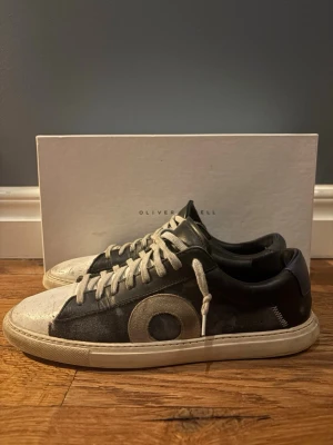 Svarta sneakers från Oliver Cabell - Snygga svarta sneakers från Oliver Cabell med vit tå och sula. Skorna har blå mocka camo detaljer   och ett stort O på sidan. De har vita skosnören och en klassisk låg profil. Materialmix av läder och mocka ger en stilren och modern look. Skorna passar upp till 41. Box och dustbag kommer med