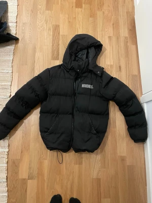 Svart pufferjacka från Hoodrich - Säljer en svart pufferjacka från Hoodrich med huva och dragkedja framtill. Jackan har två sidofickor och logga på bröstet. Perfekt för kalla dagar och riktigt skön att bära. Materialet är syntet och jackan har en klassisk pufferlook.