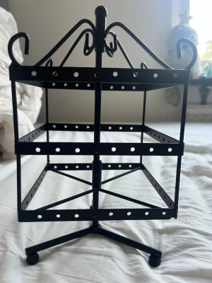 Svart roterande smyckesställ - Snyggt svart smyckesställ i metall med roterande funktion. Perfekt för att hänga örhängen, ringar eller andra småsmycken. Stället har tre våningar med hål för enkel organisering och dekorativa detaljer upptill. Passar perfekt på sminkbordet eller i garderoben.