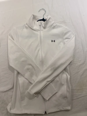 Vit ziptröja från Under Armour - Säljer en vit ziptröja från Under Armour med hög krage och hel dragkedja framtill. Tröjan har diskret logga på bröstet och är tillverkad i ett mjukt syntetmaterial som passar perfekt till träning eller som ett extra lager.