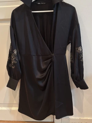 Svart omlottklänning från Zara - Snygg svart omlottklänning från Zara med v-ringning och broderade blommor på ärmarna. Klänningen har trekvartsärm, draperad detalj framtill och stängs med en dekorativ ring. Perfekt för dig som vill ha en stilren men unik look.