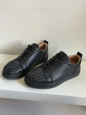 Svarta sneakers med nitar Christian Louboutin - Säljer ett par svarta sneakers i läder från Christian Louboutin. Skorna har lågt skaft och är dekorerade med svarta nitar över tån för en edgy look. Klassisk snörning och svart sula. Perfekta för dig som vill sticka ut med din stil.