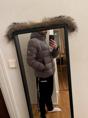 Moncler Marque Size 4 - Säljer en grå dunjacka från Moncler med huva och avtagbar pälskant. Jackan har en puffig design, dragkedja framtill och fickor på sidorna samt en ficka med logga på ärmen. Perfekt för kalla vinterdagar. Litet hål i ena innerfickan som enkelt kan sys hemma eller hos skräddare. 