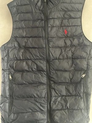 Svart dunväst från Polo Ralph Lauren - Snygg svart dunväst från Polo Ralph Lauren med quiltad design och dragkedja framtill. Västen har två fickor med dragkedja och en röd broderad logga på bröstet. Perfekt för lager-på-lager och håller dig varm med lättviktig känsla.