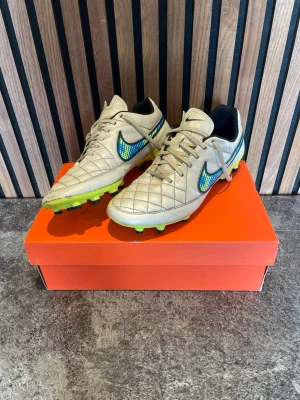 Beige Nike fotbollsskor med neon-detaljer - Snygga beige fotbollsskor från Nike med blått och gult swoosh-märke på sidorna och neongul sula. Skorna har klassisk snörning och är tillverkade i syntetmaterial med. Perfekta för dig som vill sticka ut på planen. Retro vibe