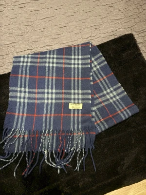 Blårutig halsduk från Burberry - Snygg halsduk från Burberry i blått med klassiskt rutmönster i vitt och rött. Halsduken har fransar i ändarna och är tillverkad i mjuk ull. Perfekt accessoar för att lyfta din outfit med en tidlös och trendig look. Den är i väldigt fint skick. Köpt på sicuro fashion!