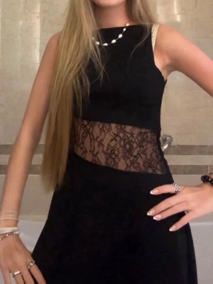 Svart klänning med spetsdetalj - Snygg svart klänning med ärmlös design och transparent spets i midjan som ger en unik look. Klänningen har en enkel, elegant form och passar perfekt för dig som vill sticka ut. Perfekt att styla med smycken och accessoarer.