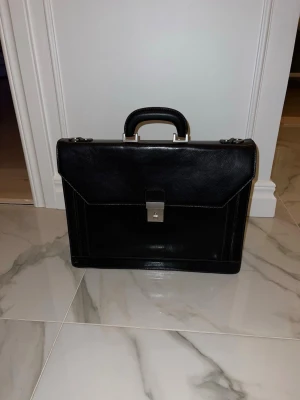 Svart portfölj i skinn med metallspänne - Stilren svart portfölj i skinn med klassisk rektangulär form och robust handtag. Väskan har ett snyggt metallspänne framtill och detaljer i silverfärgad metall. Perfekt för att bära laptop, papper och andra viktiga grejer till skolan eller jobbet.