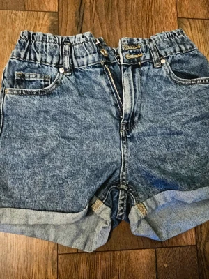 Blå denimshorts med hög midja - Snygga blå denimshorts med hög midja och elastisk resår i midjan. Shortsen har uppvikta benslut, klassiska fem fickor och dubbla knappar framtill med dragkedja. Perfekta för varma dagar och enkel att matcha med olika toppar.