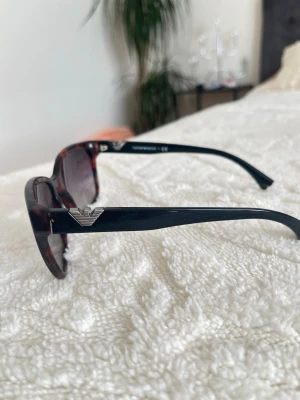 Emporio Armani bruna solglasögon - Snygga solglasögon från Emporio Armani med brunt sköldpaddsmönstrat båge och svarta skalmar. Cat-eye formade glas och silverfärgad logga på sidan. Perfekta för att lyfta din stil i sommar!