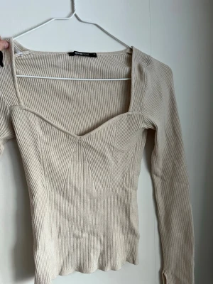 Ribbad beige långärmad topp Gina Tricot - Säljer en beige ribbad långärmad topp från Gina Tricot med snygg v-ringning och figurnära passform. Toppen har dekorativa sömmar framtill och är gjord i ett stretchigt material som sitter bekvämt. Perfekt för att skapa en trendig look. Storlek XS