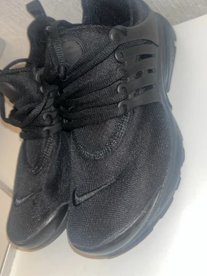 Svarta Nike Air Presto sneakers - Snygga helsvarta Nike Air Presto sneakers med mesh-ovandel och plastdetaljer vid snörningen. Skorna har en låg profil och en bekväm, flexibel sula som passar perfekt till en sportig och urban stil. Diskret Nike-logga på sidan och tungan.