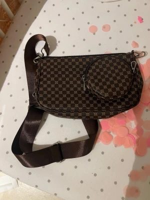 Brun axelväska med monogrammönster - Snygg brun axelväska med klassiskt monogrammönster i beige. Väskan har en bred justerbar axelrem och en rund liten ficka framtill. Perfekt för att bära det viktigaste med stil.