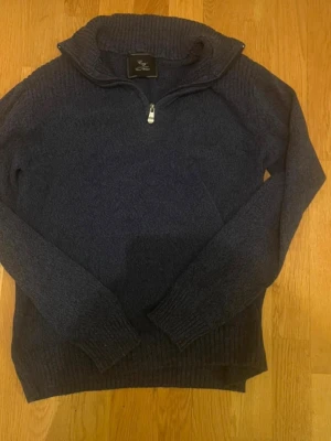 Mörkblå stickad tröja med halvzip - Mörkblå stickad tröja från Gant med halvzip och hög krage. Tröjan har långa ärmar och ribbade muddar vid ärmslut och nederkant. Perfekt för lager på lager och en klassisk stil. Materialet känns mjukt och värmande.
